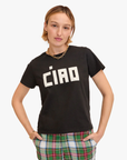 A person with short, light brown hair is wearing a black Clare Vivier Classic Tee made from 100% cotton, featuring the word "Ciao" in bold, white letters. They have gold hoop earrings, a gold chain necklace, and gold bracelets. Additionally, they are wearing plaid pants with green, red, and white stripes.