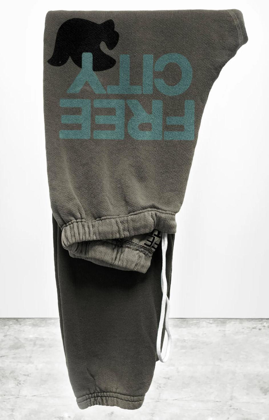 The SUPERFLUFF Lux OG Sweatpant by Free City (sparrow, LLC) is a pair of gray unisex sweatpants, folded and draped over a light gray, cement-like surface. These pants feature a graphic of a black bear above the turquoise text "FREE CITY" on one leg and are accented with white drawstrings. Made from lightweight French terry, they boast hand-screen printed designs done locally for a unique charm.