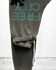 The SUPERFLUFF Lux OG Sweatpant by Free City (sparrow, LLC) is a pair of gray unisex sweatpants, folded and draped over a light gray, cement-like surface. These pants feature a graphic of a black bear above the turquoise text "FREE CITY" on one leg and are accented with white drawstrings. Made from lightweight French terry, they boast hand-screen printed designs done locally for a unique charm.