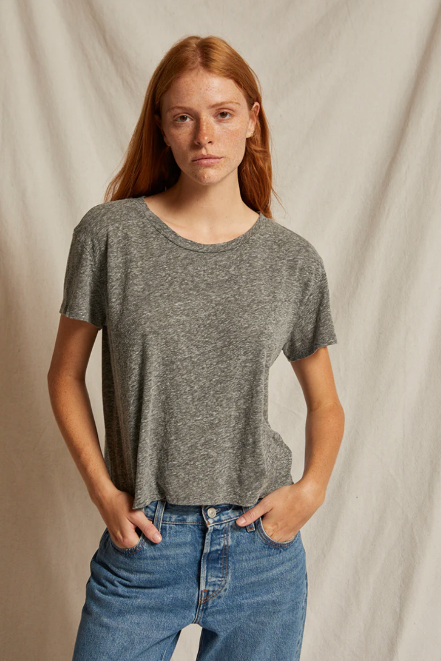 A person with long red hair and freckles is wearing a grey Harley SS Boxy Crew from Perfectwhitetee and blue jeans in a casual style. They have their hands in their pockets, standing against a light-colored, textured backdrop that evokes the warm hues of Arizona, looking directly at the camera with a neutral expression.