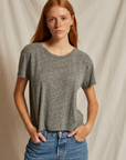 A person with long red hair and freckles is wearing a grey Harley SS Boxy Crew from Perfectwhitetee and blue jeans in a casual style. They have their hands in their pockets, standing against a light-colored, textured backdrop that evokes the warm hues of Arizona, looking directly at the camera with a neutral expression.