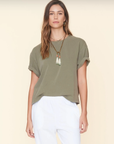 A person with long, brown hair wears a loose-fitting olive green Palmer Tee by Xirena with rolled sleeves, a long beaded necklace with white and green tassels, and white pants. The ultra-soft cotton jersey shirt accentuates their relaxed posture as they stand against a neutral background, looking straight ahead.