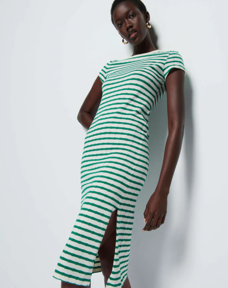 A woman is wearing a verdant green stripe, 100% Pima cotton, short-sleeved Ingrid Shrunken T-Shirt Dress by Nation LTD with a high slit. She stands against a plain background, tilting her head slightly and looking towards the camera. Her outfit is complemented by large gold earrings, making it an essential t-shirt dress for any wardrobe.