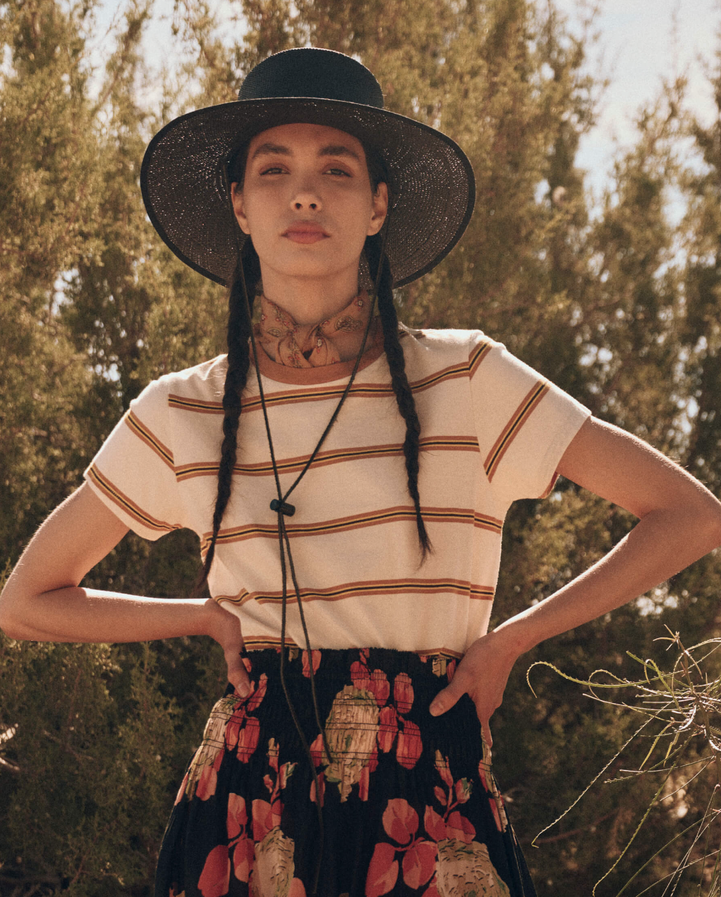 A young woman wearing a black wide-brimmed hat and patterned scarf stands confidently outdoors in her The Little Tee by The Great Inc. She is dressed in a striped white t-shirt and a floral skirt, with her hair styled in two long braids. The background features trees and a clear sky.