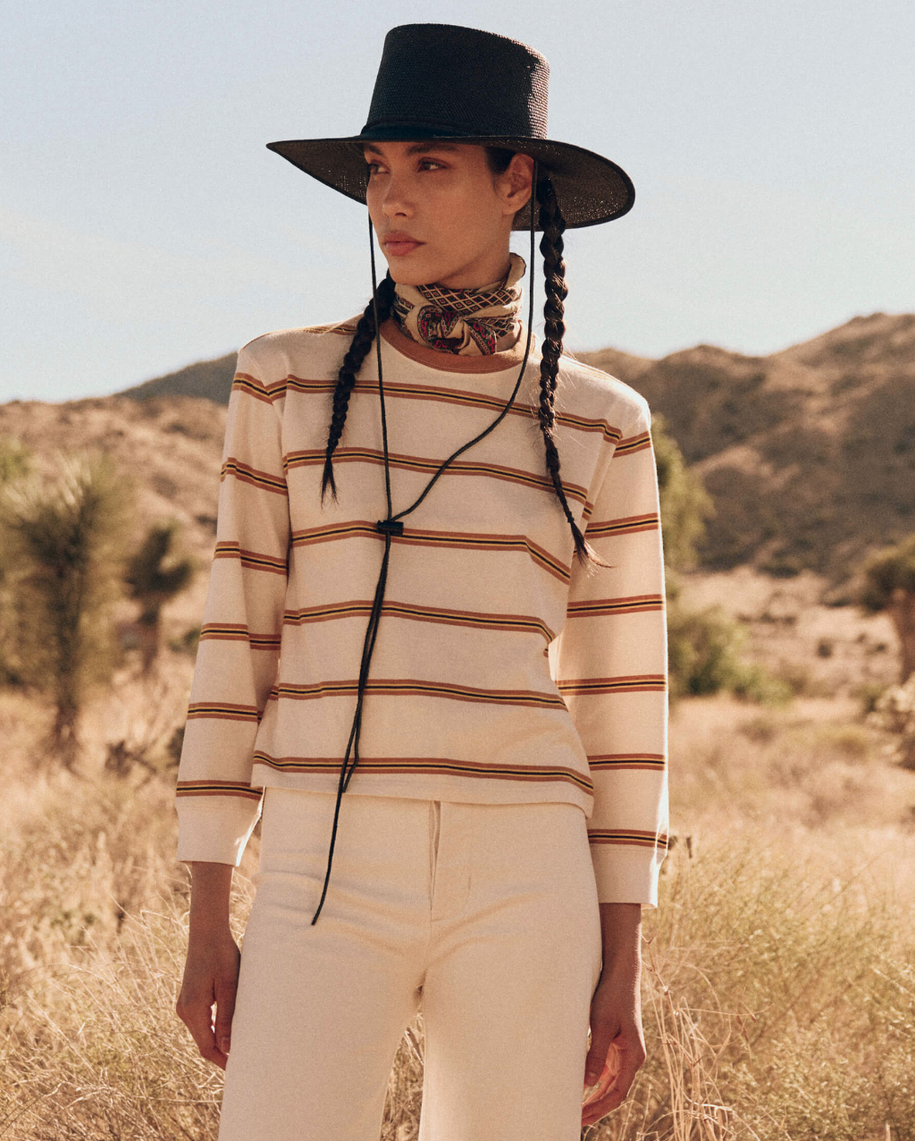 A person stands in a desert landscape wearing a broad-brimmed black hat with chin straps, The Campus Crew by The Great Inc., and light-colored pants. The individual has two braids and wears a patterned scarf around their neck. In the background, there are desert plants and hills.