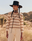 A person stands in a desert landscape wearing a broad-brimmed black hat with chin straps, The Campus Crew by The Great Inc., and light-colored pants. The individual has two braids and wears a patterned scarf around their neck. In the background, there are desert plants and hills.