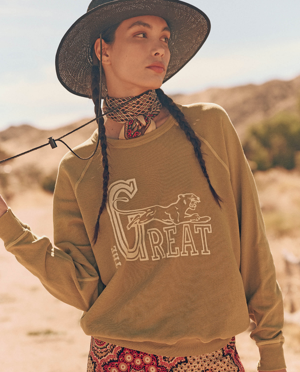 A person stands outdoors in a desert landscape, wearing a wide-brimmed hat, a bandana around their neck, and The College Sweatshirt by The Great Inc., handcrafted in Los Angeles with the word &quot;GREAT&quot; and a wild panther graphic. The person looks to the side, while their other hand lightly holds the hat&#39;s strap.