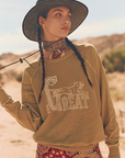 A person stands outdoors in a desert landscape, wearing a wide-brimmed hat, a bandana around their neck, and The College Sweatshirt by The Great Inc., handcrafted in Los Angeles with the word "GREAT" and a wild panther graphic. The person looks to the side, while their other hand lightly holds the hat's strap.