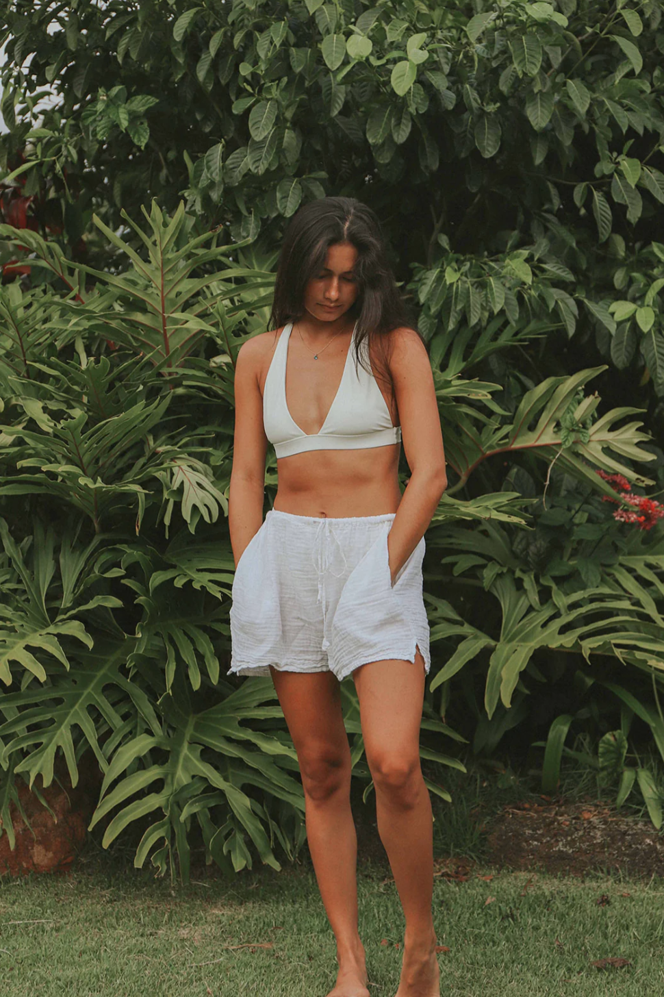 A woman with long dark hair stands barefoot in front of lush greenery. She is wearing a white bra-style top and cute Hawaiian Beach Short by By the Sea Organics with a drawstring waist, her hands tucked into the pockets. She gazes downward with a calm expression, exuding effortless elegance.