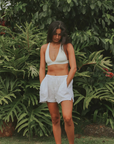 A woman with long dark hair stands barefoot in front of lush greenery. She is wearing a white bra-style top and cute Hawaiian Beach Short by By the Sea Organics with a drawstring waist, her hands tucked into the pockets. She gazes downward with a calm expression, exuding effortless elegance.