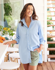 A woman in a 100% cotton, oversized SHIRLEY Shirt by Frank & Eileen and light green shorts stands in a bright kitchen. She has one hand in her pocket and smiles while leaning on the counter. Her everyday go-to outfit complements the oranges, yellow flowers, and potted plants around her.