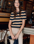 A person with long hair is standing and smiling in a cozy café named The Brentwood. They are wearing a striped navy blue and beige Modern Tee by Kule, tucked into high-waisted denim, while holding a dark jacket. The café has a warm ambiance with French menu items written on the walls.