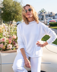 A person with wavy blonde hair wearing sunglasses poses casually beside a vintage white convertible car. They are dressed in the luxurious Monterey Sweater, knitted by artisans from fine Italian cotton by Frank & Eileen, along with matching pants. A bouquet of soft pink and white flowers is placed on the car's trunk.