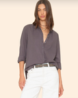 A woman with long, brown hair wears the Beau Shirt from Xirena, a dark gray, menswear-inspired button-up crafted from cotton poplin. She has the sleeves rolled up and pairs it with white pants. Layered necklaces adorn her as she stands against a light beige background.