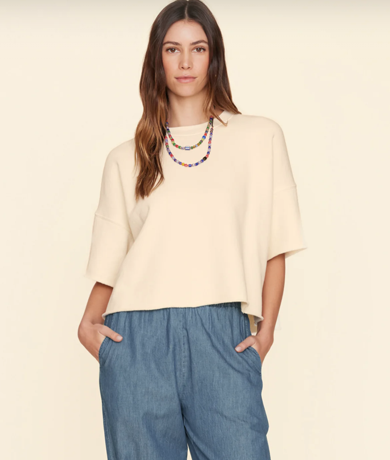 A woman with long brown hair wears a loose, cream-colored Romeo Sweatshirt by Xirena and blue high-waisted pants. She has her hands in her pockets and is adorned with a colorful beaded necklace. The background is a plain off-white color, highlighting her elevated comfort.