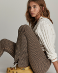 A woman with long, wavy hair is seated against a plain background. She wears a white knit sweater, LIBERTINE FLORENCE FLARE high-waisted patterned brown pants by The Upside, and yellow clogs with decorative charms. Her expression is neutral, and she looks slightly to the side.