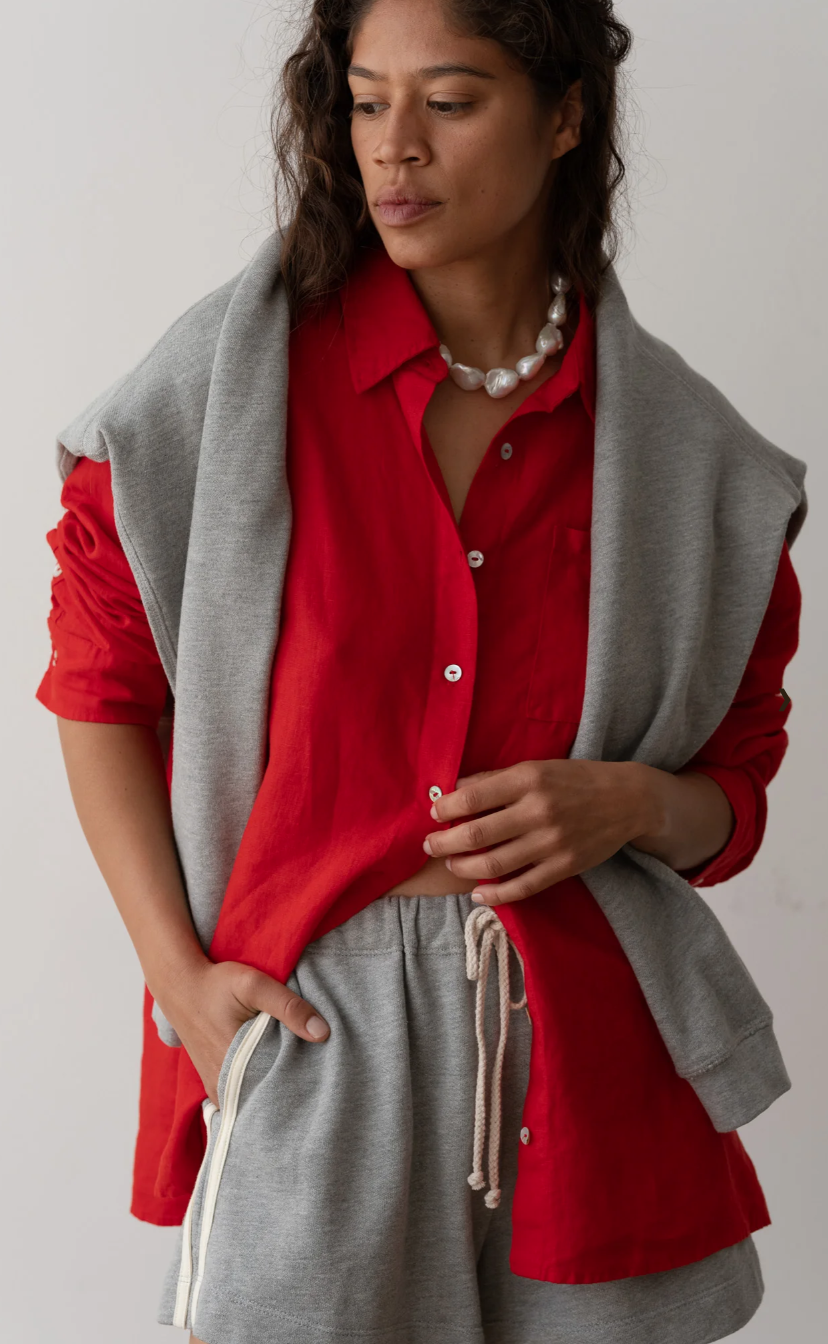 A person with wavy hair wears The Linen Relaxed Shirt in a bright red color by Donni, paired with a grey sweatshirt draped over their shoulders and grey drawstring shorts. They accessorize with a chunky pearl necklace and have a contemplative expression while posing with one hand in their pocket.