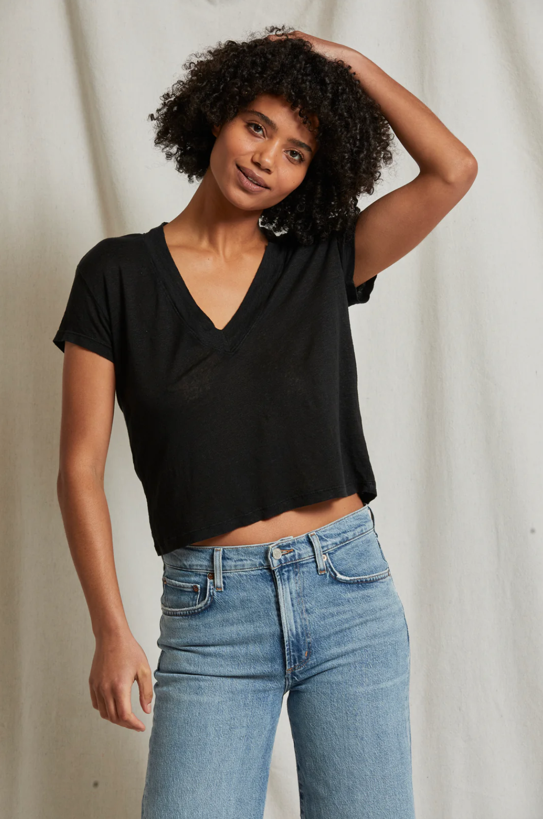 A person with curly hair stands against a light-colored backdrop, wearing the Rosie V-neck short-sleeve top from Perfectwhitetee and blue jeans. They have one hand behind their head and are looking at the camera with a relaxed expression.