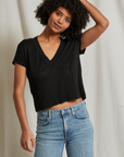 A person with curly hair stands against a light-colored backdrop, wearing the Rosie V-neck short-sleeve top from Perfectwhitetee and blue jeans. They have one hand behind their head and are looking at the camera with a relaxed expression.