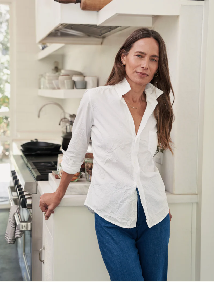 A woman with long hair stands casually in a kitchen, leaning against the counter. She is wearing a white BARRY CRINKLE tailored-fit button-down shirt from Frank & Eileen, made from 100% cotton, along with blue jeans. Behind her, there is a stove with cookware and shelves filled with various kitchen items. The kitchen has a bright and airy feel.