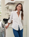 A woman with long hair stands casually in a kitchen, leaning against the counter. She is wearing a white BARRY CRINKLE tailored-fit button-down shirt from Frank & Eileen, made from 100% cotton, along with blue jeans. Behind her, there is a stove with cookware and shelves filled with various kitchen items. The kitchen has a bright and airy feel.