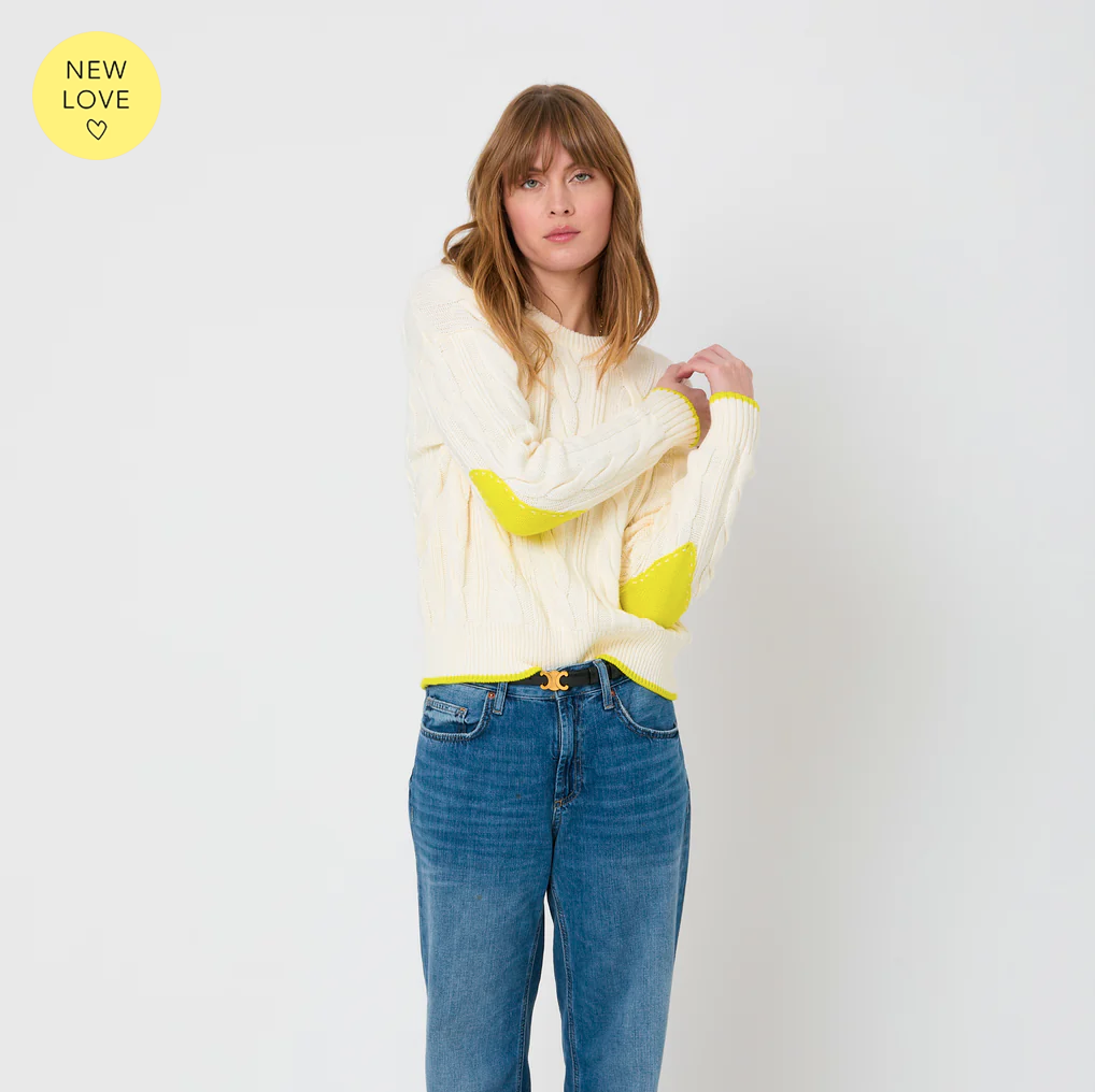 A woman with long brown hair is wearing the cozy Maxwork Cable Sweater by Kerri Rosenthal, made from a comfortable cotton blend in a cream color, featuring stylish yellow accents on the elbows and hem. She pairs it with blue jeans and stands against a plain white background. In the top left corner, there's a yellow circle with the text "New Love" and a heart symbol, making it perfect for fall season fashion.