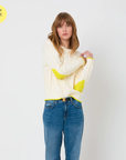 A woman with long brown hair is wearing the cozy Maxwork Cable Sweater by Kerri Rosenthal, made from a comfortable cotton blend in a cream color, featuring stylish yellow accents on the elbows and hem. She pairs it with blue jeans and stands against a plain white background. In the top left corner, there's a yellow circle with the text "New Love" and a heart symbol, making it perfect for fall season fashion.