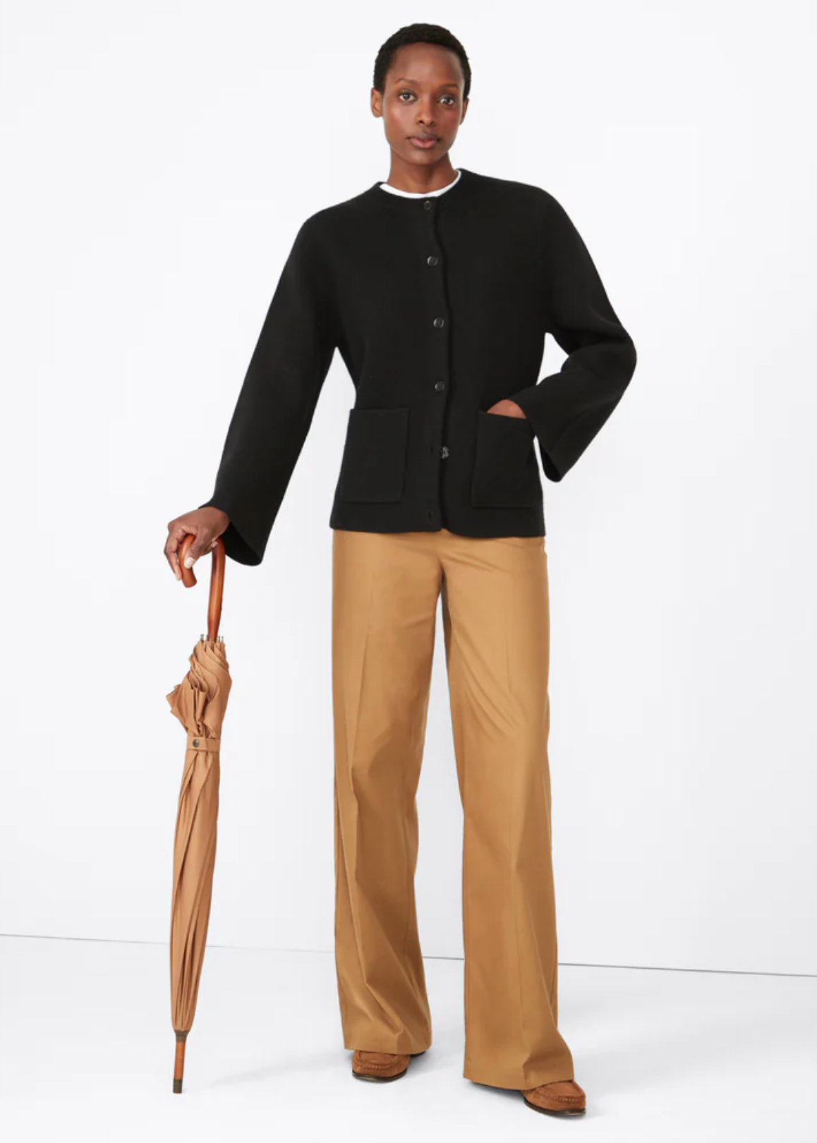 A person stands against a plain white background, wearing "The Marta" black button-up jacket with a flattering fit from Kule, khaki wide-leg pants, and brown shoes. They hold a closed tan umbrella in their right hand.