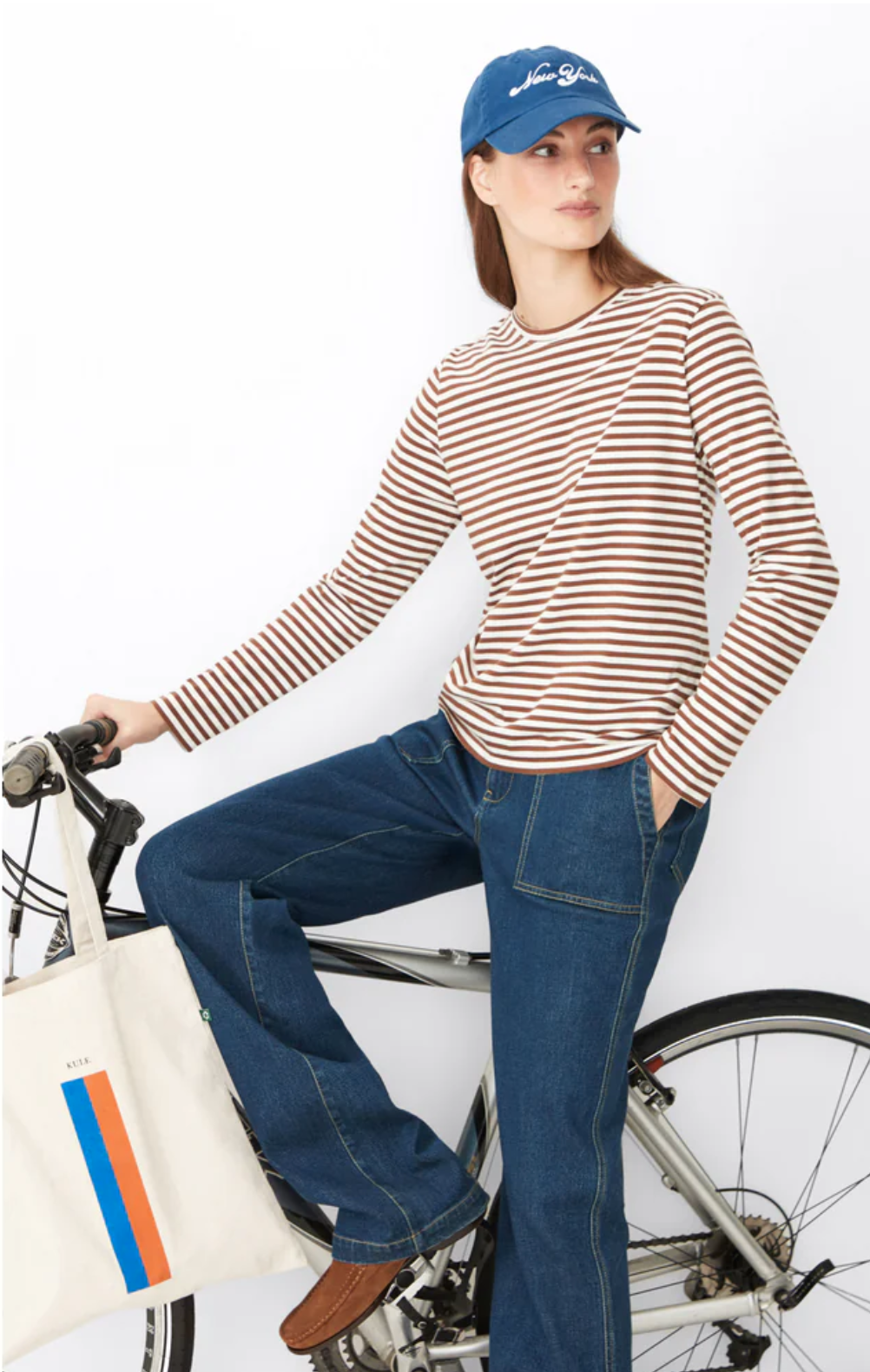 A woman wearing a blue baseball cap, The Modern Long by Kule in brown and white stripes, and blue jeans is standing beside a bicycle. She has one foot on a pedal and is holding a tote bag with a colorful striped design over the handlebars.