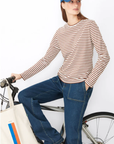 A woman wearing a blue baseball cap, The Modern Long by Kule in brown and white stripes, and blue jeans is standing beside a bicycle. She has one foot on a pedal and is holding a tote bag with a colorful striped design over the handlebars.