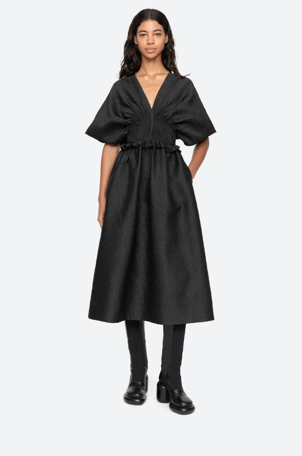 A woman stands against a plain background wearing a Sea New York Elegi Solid Short Sleeve Dress with oversized sleeves and a deep V-neckline. The dress has a cinched waist and an A-line skirt that reaches below the knees. She pairs it with black boots and has long, wavy hair.