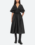 A woman stands against a plain background wearing a Sea New York Elegi Solid Short Sleeve Dress with oversized sleeves and a deep V-neckline. The dress has a cinched waist and an A-line skirt that reaches below the knees. She pairs it with black boots and has long, wavy hair.