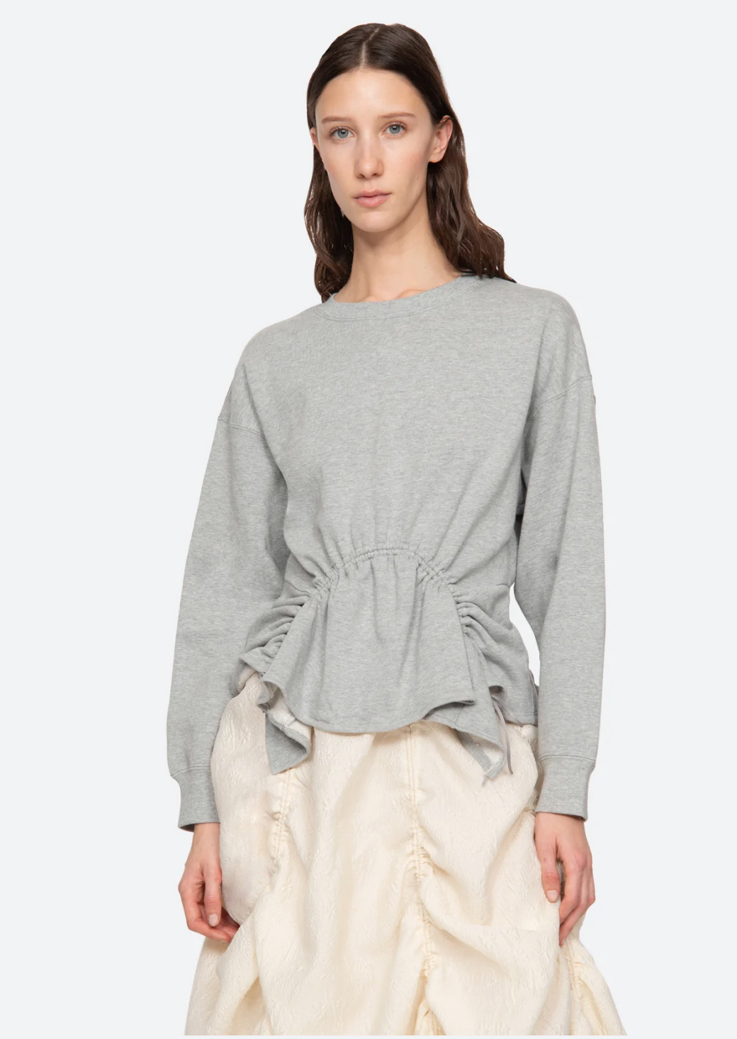 A person with long, dark hair is wearing the Elegi Solid Long Sleeve Sweatshirt by Sea New York, a light gray top with a cinched waist design, paired with a cream-colored, textured skirt featuring an asymmetrical hem. They are standing against a plain white background and looking directly at the camera.