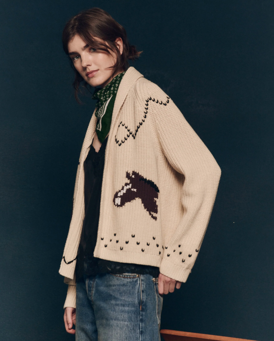 A person stands against a dark background wearing the beige Horse Lodge Cardigan from The Great Inc., crafted from cotton blend yarn and adorned with hand-embroidered horse head and whip details. They pair it with a black top, blue jeans, and a green bandana around their neck. With short, dark hair, they gaze slightly to the side.