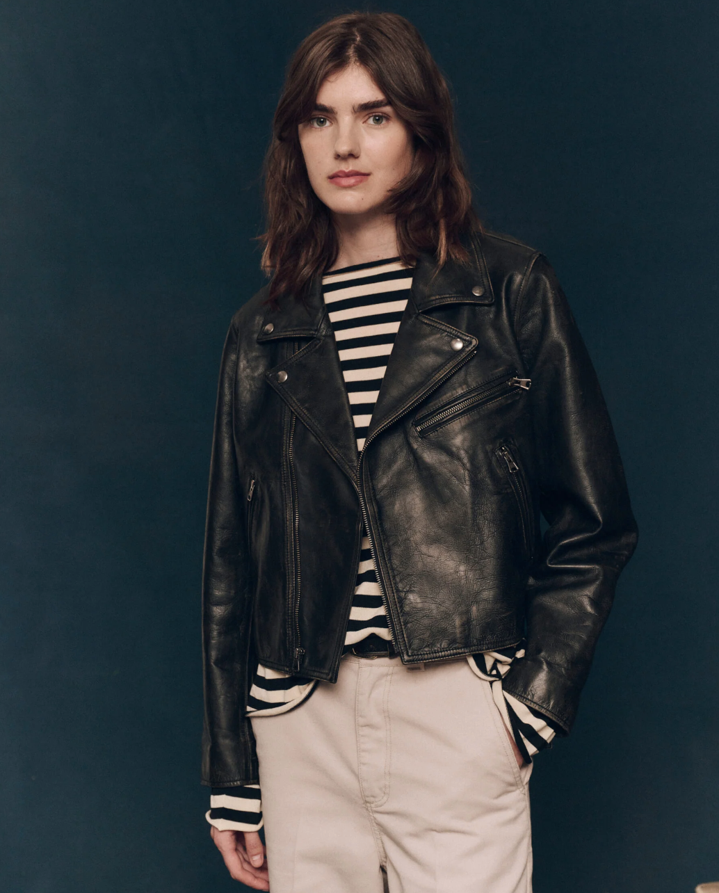 A person with shoulder-length brown hair is wearing The Perfect Leather Jacket by The Great Inc. over a black and white striped shirt. They are posing against a dark blue background, with their hands in the pockets of their light-colored pants adorned with antique silver snaps.