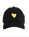 A black baseball cap from Kerri Rosenthal's KR CAP-Sule Collection, named the Embroidered Heart Hat, featuring a small yellow heart embroidered on the front center. This collegiate hat has a curved brim, visible stitching, and an adjustable back strap. The background is plain white.