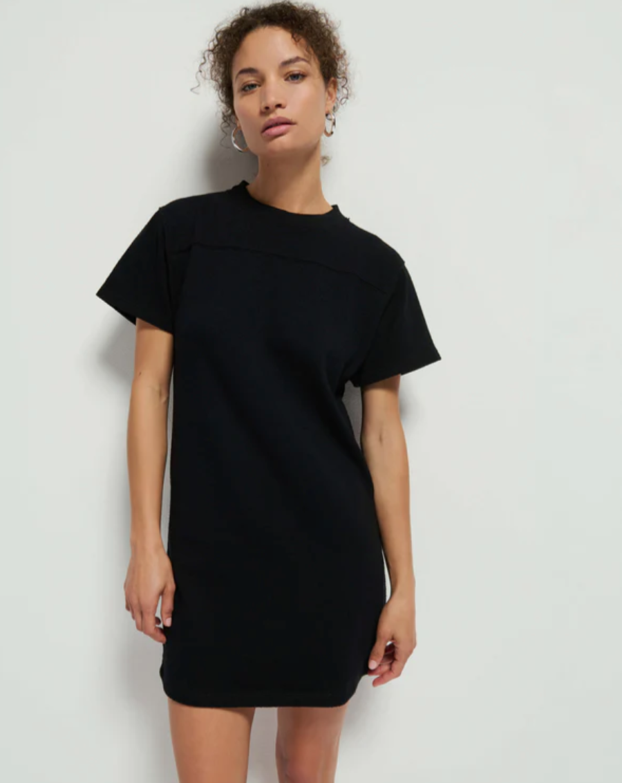 A person with curly hair stands against a white background, wearing a short-sleeved Finn Crewneck Dress in Jet Black from Nation LTD. They have a relaxed posture with one hand hanging by their side and the other hand resting on their thigh.