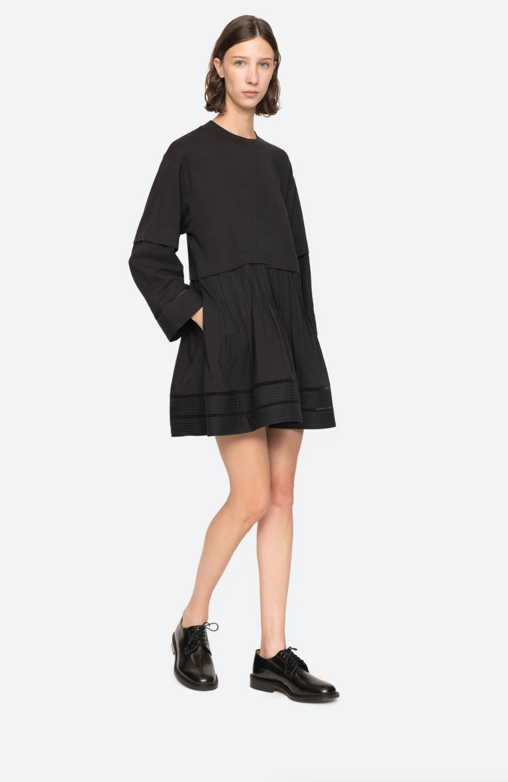 A person with shoulder-length brown hair is standing against a plain background wearing the Sea New York Renata Tunic Dress, which features a relaxed fit, long sleeves, and a tiered skirt. They are also wearing black lace-up shoes and are looking towards the camera with a neutral expression.