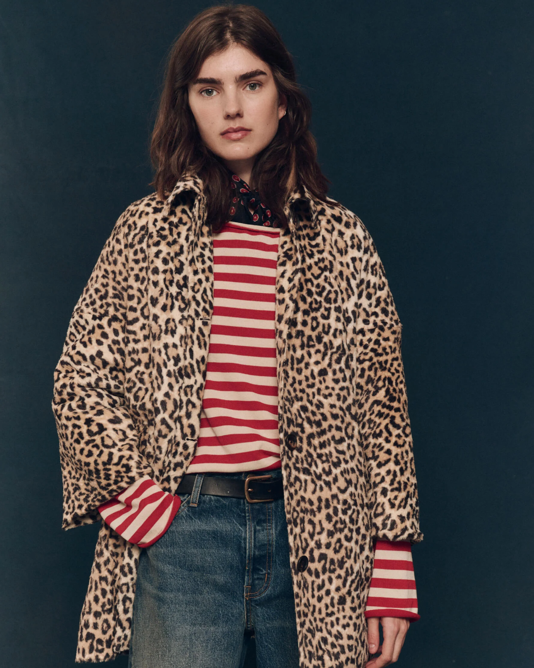 A woman stands against a dark background, wearing The Car Coat from The Great Inc. in a stylish leopard print over a red and white striped shirt. She has long, dark hair and a neutral expression. She is also wearing blue jeans with one hand in her pocket.