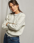 A person with long hair stands with arms crossed, wearing the Ziggy Inside Out LS Crewneck Sweatshirt from Perfectwhitetee in light gray and blue jeans. They are looking directly at the camera with a neutral expression. The background is plain and light-colored.