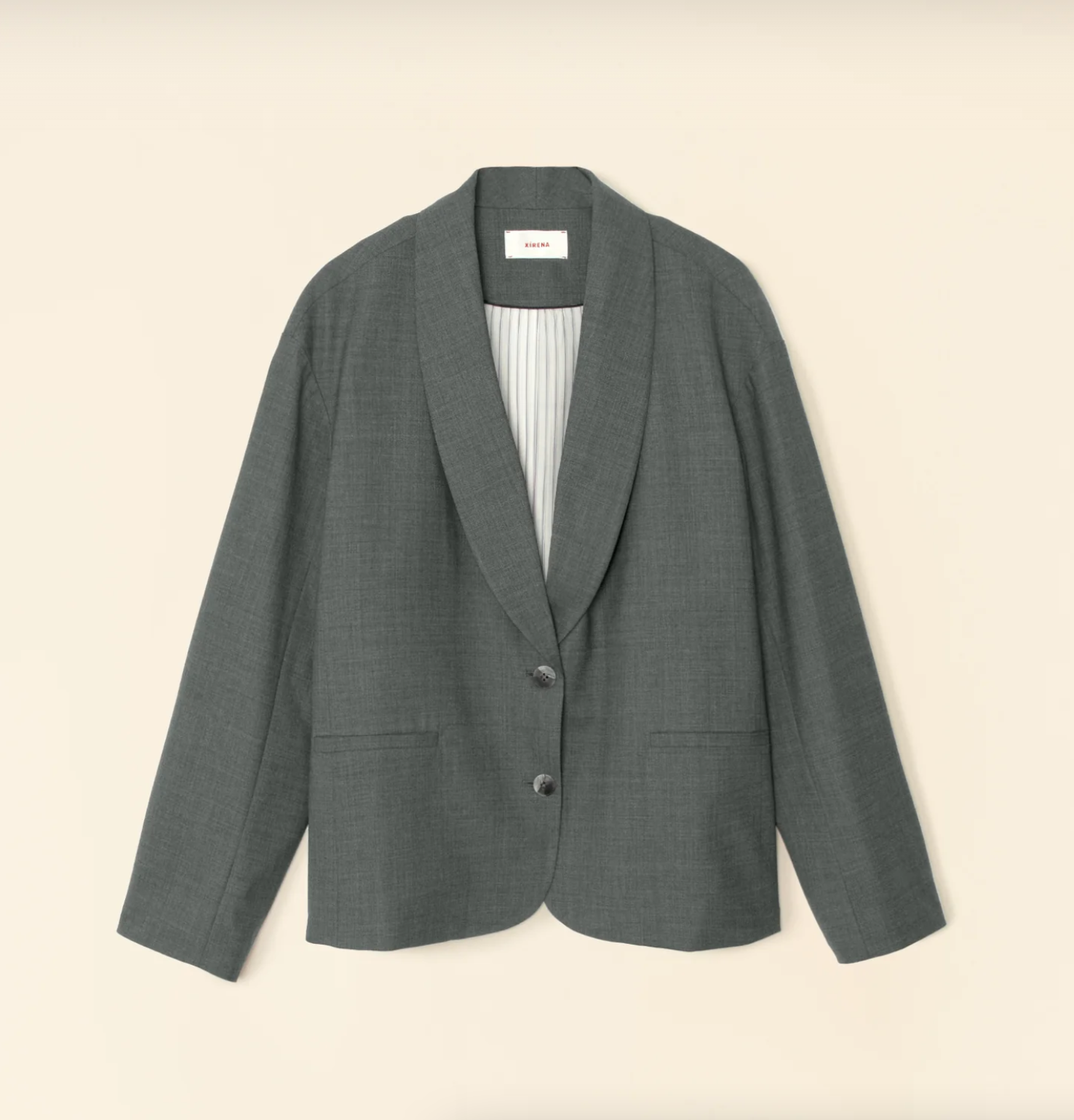 The Jonah Jacket by Xirena, a gray wool blazer with long sleeves and a relaxed, unstructured fit, hangs against a beige background. It features a shawl collar, two front buttons, and two front pockets. The interior lining is accentuated with contrast color piping that highlights a subtle stripe pattern for a soft-tailored look.