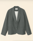 The Jonah Jacket by Xirena, a gray wool blazer with long sleeves and a relaxed, unstructured fit, hangs against a beige background. It features a shawl collar, two front buttons, and two front pockets. The interior lining is accentuated with contrast color piping that highlights a subtle stripe pattern for a soft-tailored look.