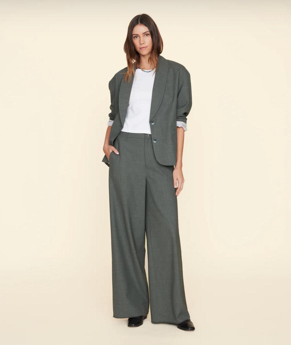 A woman stands against a plain background wearing a loose-fitting grey pantsuit with wide-legged, wool blend trousers and a matching blazer over a white t-shirt. She has long brown hair and is wearing black shoes. Her left hand is in her pants pocket, showcasing the frayed raw hem of her stylish Portlyn Pant by Xirena.