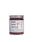 A jar of Faire Sichuan Spicy Dan Dan Sauce with a silver lid and a white label. The label features text in English and Chinese characters, along with the image of a red chili pepper and a white hand holding a pair of chopsticks, promising rich umami flavor perfect for any Chinese noodle dish.