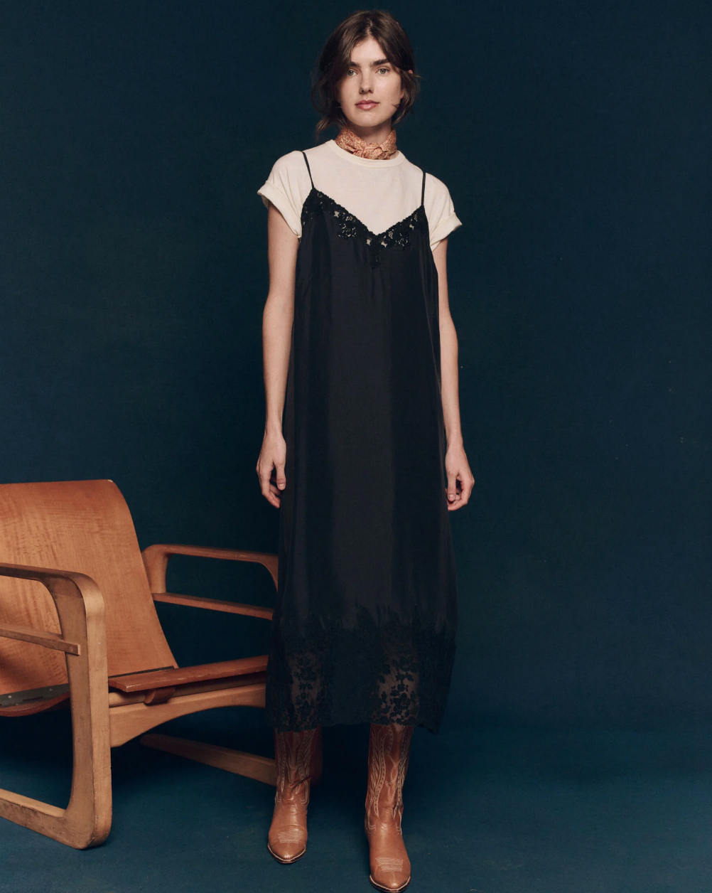 A person with shoulder-length brown hair stands indoors against a dark backdrop. They're wearing a white t-shirt underneath The Legacy Dress by The Great Inc., a long black slip dress featuring adjustable shoulder straps and detailed with intricately designed embroidered mesh lace. They’ve paired this ensemble with light brown cowboy boots, standing beside a wooden chair.