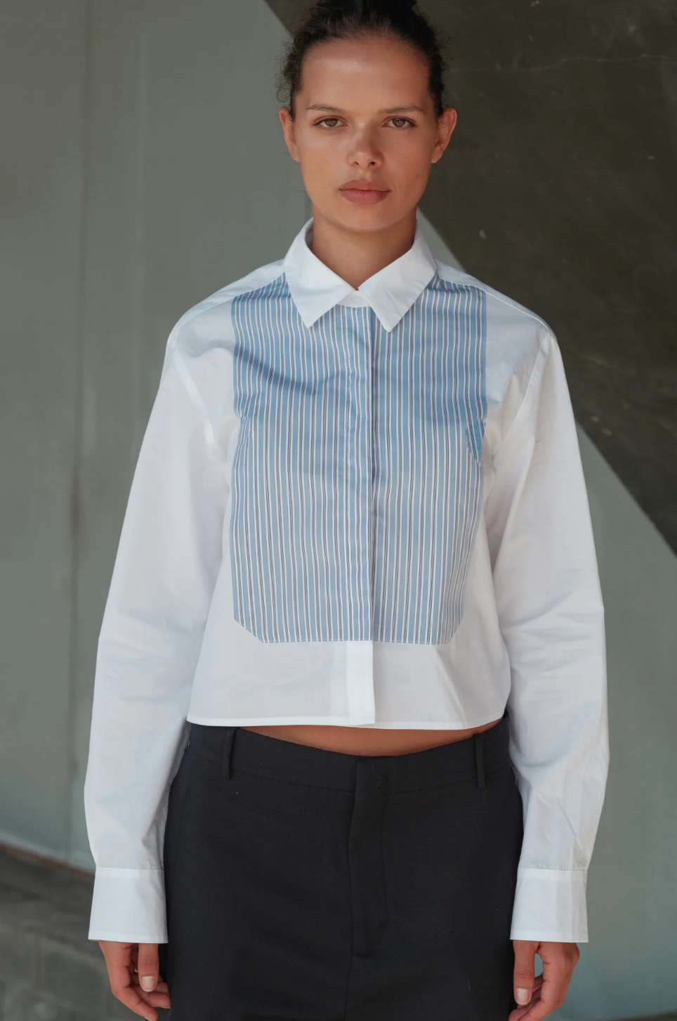 A person with dark hair pulled back stands facing the camera, wearing The BIB Shirt by Cissa, made from premium Egyptian cotton. This contemporary shirt features a formal white design with a light blue vertical stripe panel and is paired with black trousers. The background is neutral, presented in shades of gray with a slight diagonal element.