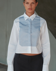 A person with dark hair pulled back stands facing the camera, wearing The BIB Shirt by Cissa, made from premium Egyptian cotton. This contemporary shirt features a formal white design with a light blue vertical stripe panel and is paired with black trousers. The background is neutral, presented in shades of gray with a slight diagonal element.