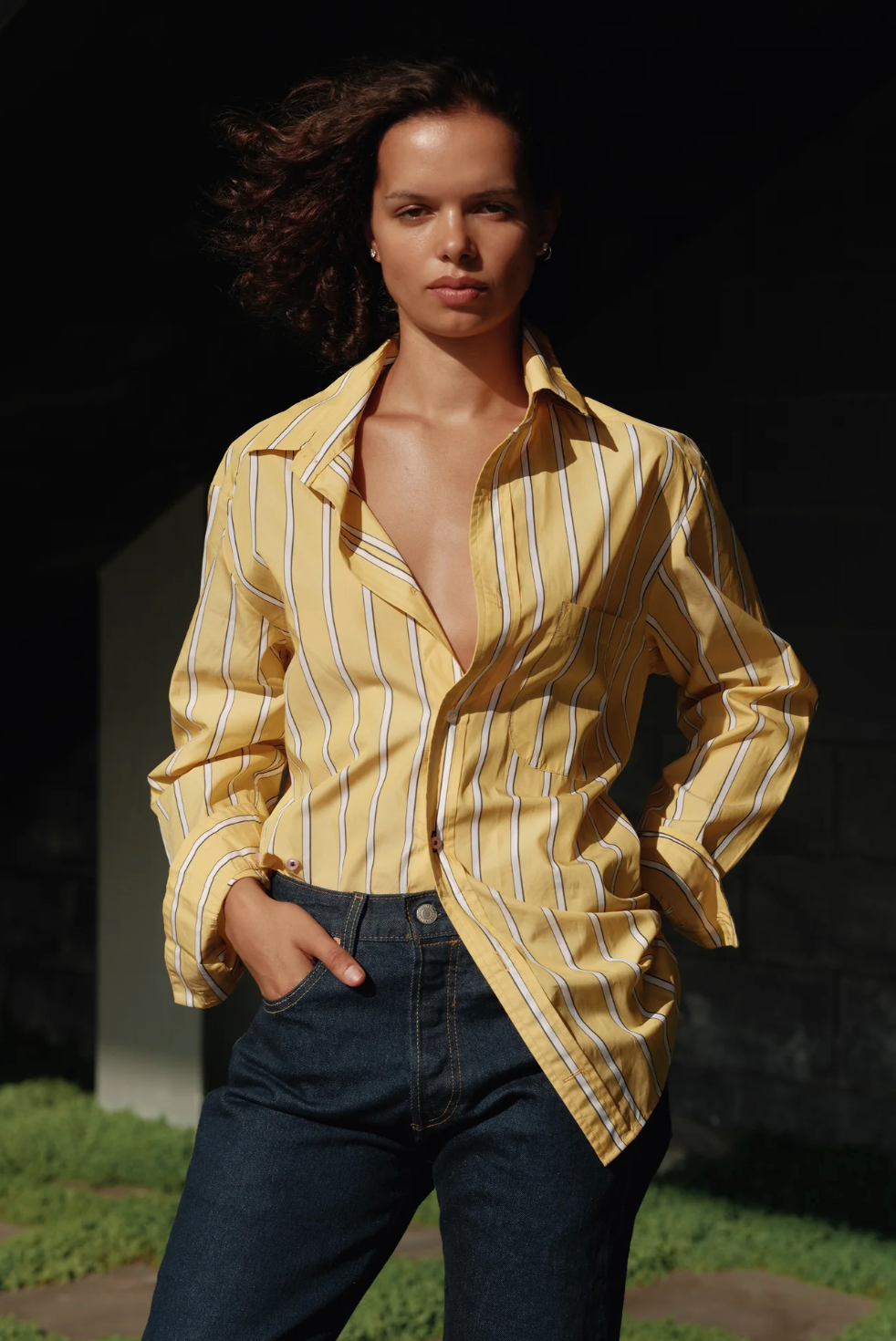 A person with shoulder-length wavy hair stands outdoors in sunlight, wearing The MEN'S Shirt by Cissa, a yellow and white striped men's shirt made from soft Egyptian cotton, with the top buttons undone, and dark blue jeans. Their hands are in their pockets, and they have a confident expression.