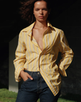 A person with shoulder-length wavy hair stands outdoors in sunlight, wearing The MEN'S Shirt by Cissa, a yellow and white striped men's shirt made from soft Egyptian cotton, with the top buttons undone, and dark blue jeans. Their hands are in their pockets, and they have a confident expression.