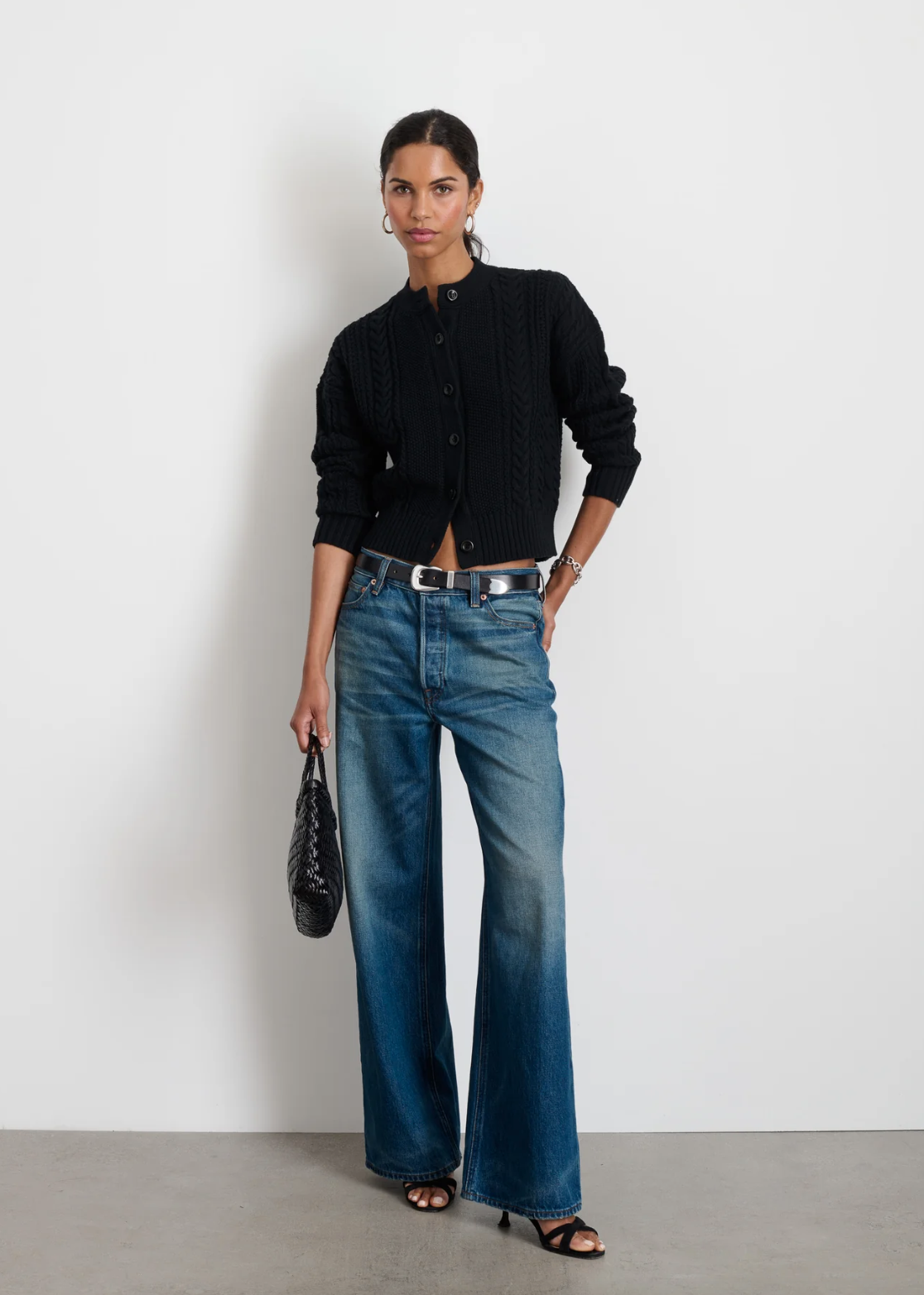 A person stands against a white wall, wearing the Alex Mill Cable Knit Nico Chunky Cardigan in black and blue high-waisted, wide-leg jeans. They hold a black woven handbag and wear black heeled sandals. Their hair is pulled back, and they pose with one hand in their pocket.